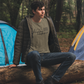Happy Camper T-Shirt in Heather Olive. The front has a cursive font "happy" with "camper" below it in a bold lowercase font. Two triangle mountains are above the word happy, and three pine trees are above the word camper. The design is in black. This is the front view of the shirt on a male model with a green coat on sitting on a large log in a woodsy area with tents behind him.
