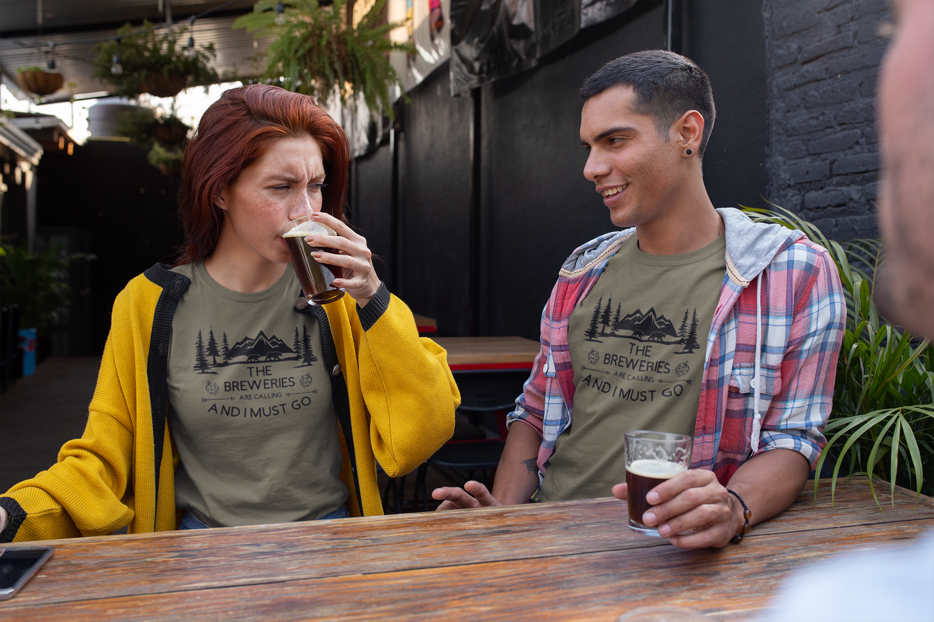 The Breweries Are Calling And I Must Go - T-Shirt in a heather Olive Garden. Black Design on the front with the saying "the breweries are calling and I must go" with arrows pointing at "are calling" hoops surrounding the "breweries" and pine trees, mountains and 2 black bears above the saying. This is the front view of the shirt on two different models male and female having a beer at a brewery.