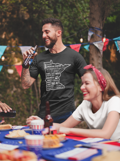 Guy model at a party drinking a beer, wearing a black Minnesota Beers - T-Shirt with hazy ipa, American porter, Oktoberfest, lager, scotch ale, stout, dunkel, saison, wheat, cream ale, Irish red ale, pilsner, pale ale, porter, kolsch, amber ale, cream ale on the front in the state of Minnesota with icons of a hop, glass of beer, barrel of beer, bottle opener, and a wheat strand.